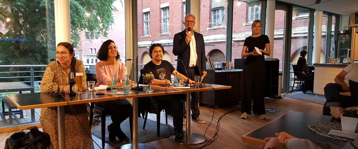 An einem Tisch sitzen die Autoren Ronya Othmann und Dan Thy Nguyen. Moderationbegrüßt die Zuschauer.