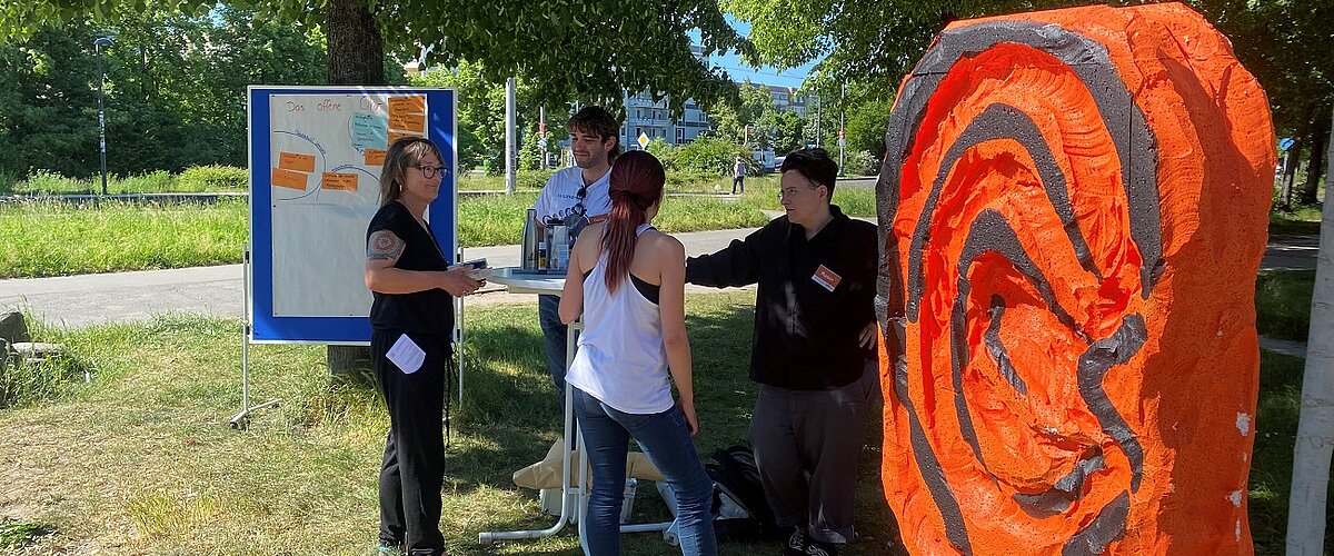 Vier Leute sprechen an einem Stehtisch neben dem orangenen plastischen Ohr