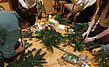 An einem Tisch werden Weihnachtskränze gebunden. Alle Utensilien und Tannenzweige liegen bereit.