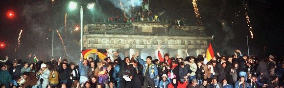 feiernde, jubelnde Menschen stehen vorm Brandenburger Tor, Feuerwerk fliegt in den Nachthimmel 