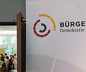 Die Tür steht offen. Im Raum sieht man viele Personen versammelt. Auf einem Banner vor der Tür steht: "Bürgerrat"