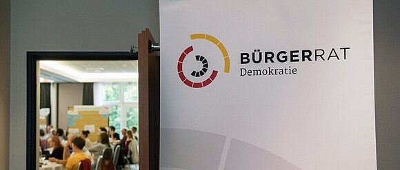 Die Tür steht offen. Im Raum sieht man viele Personen versammelt. Auf einem Banner vor der Tür steht: "Bürgerrat"