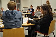 mehrere Gruppen an Tischen, Teilnehmende hören einer dozierenden Person zu, einige notieren etwas