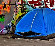 vor einer mit Graffity besprühten Seinmauer ist das blaues Zelt  eines Obdachlosen aufgebaut.