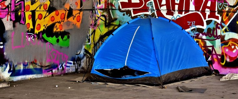 vor einer mit Graffity besprühten Seinmauer ist das blaues Zelt  eines Obdachlosen aufgebaut.