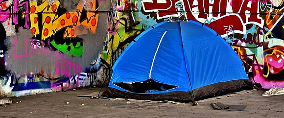 vor einer mit Graffity besprühten Seinmauer ist das blaues Zelt  eines Obdachlosen aufgebaut.