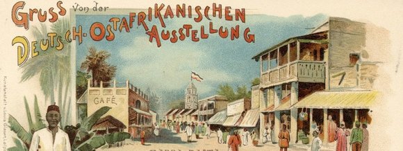 altes Postkartenmotiv: große Messeausstellungspassage mit Afrikanern in traditioneller Kleidung. Text auf der Karte: Gruß von der Deutsch-Ostafrikanischen Ausstellung