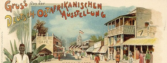 altes Postkartenmotiv: große Messeausstellungspassage mit Afrikanern in traditioneller Kleidung. Text auf der Karte: Gruß von der Deutsch-Ostafrikanischen Ausstellung